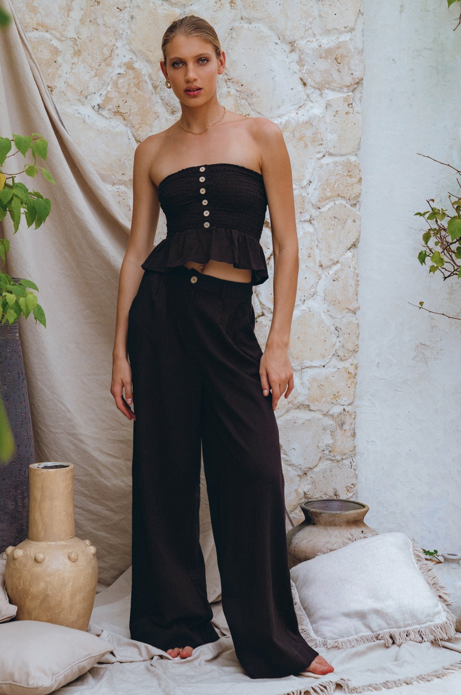 Woman wearing a black strapless top ad wide-leg Black Classic Linen Pants against a stone wall. Linen 