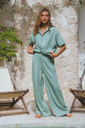 Woman wearing Sage Green Classic Linen Pants standing by a stone wall and poolside.
