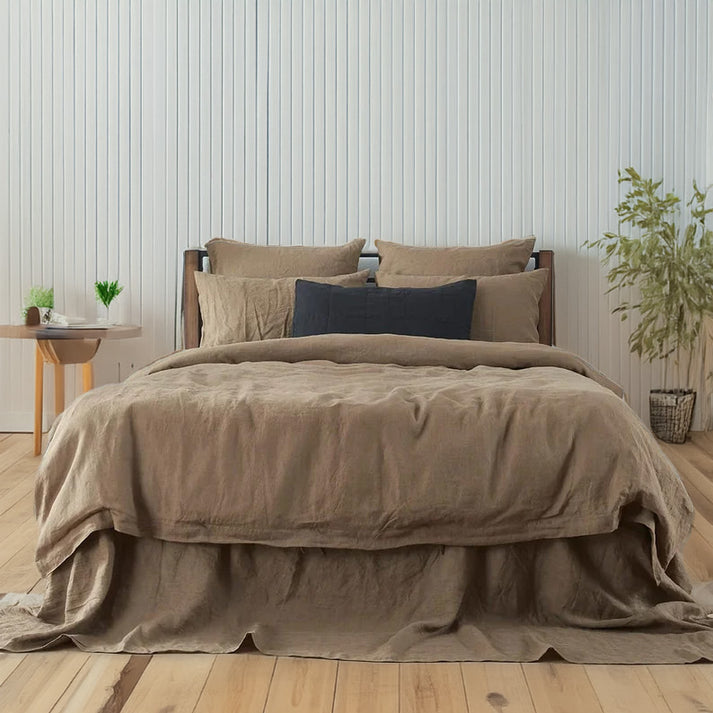 Bed with dusty brown linen and cotton bedding and pillows in a room with wooden flooring and plants.