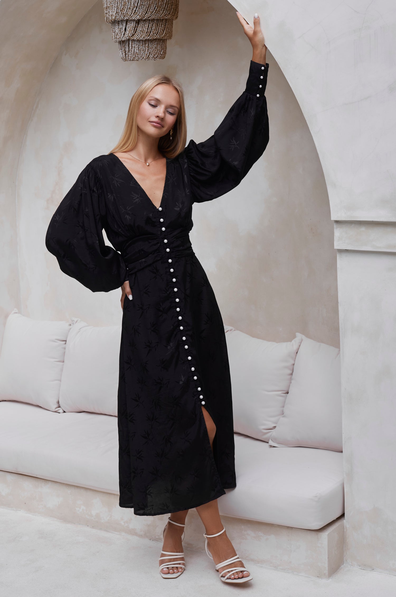 Liliana button front midi dress in black on model standing in a modern interior setting with a white sofa and decorative elements.
