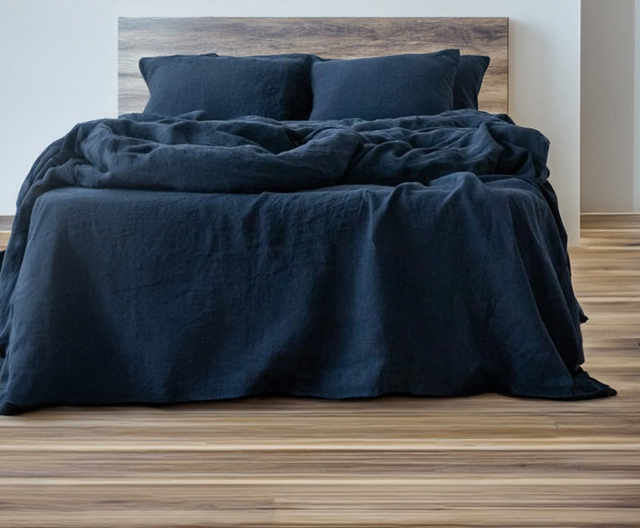 Bed with deep navy linen and cotton duvet set and wooden headboard on a wooden floor.