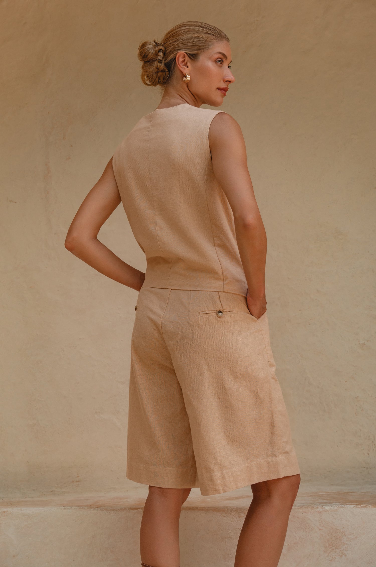 Woman wearing Riviera Linen Beige Vest and shorts back view against a beige wall.